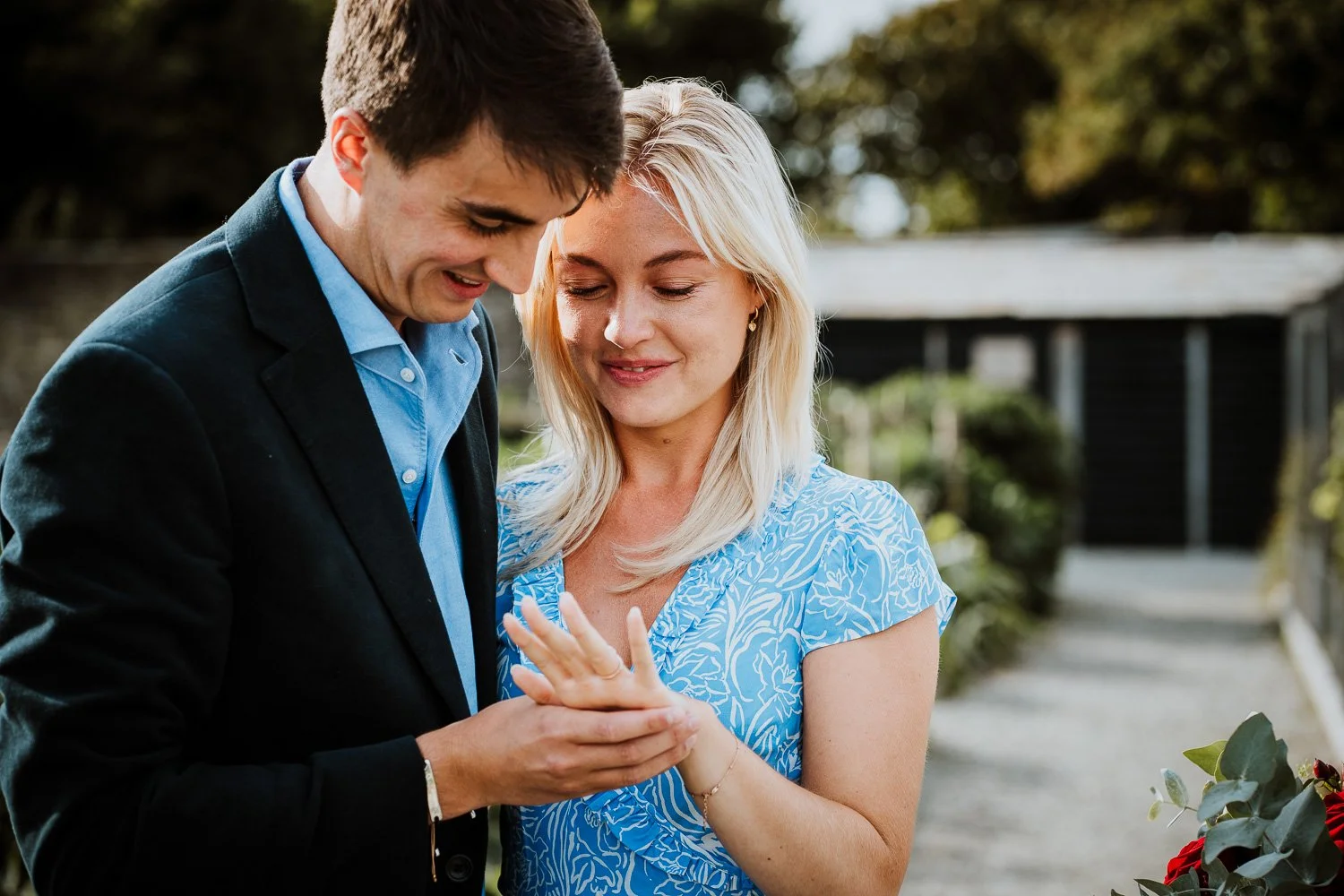 A young man proposing marriage to a woman outdoors, with both smiling and holding hands, surrounded by greenery and flowers. Natural Proposal Photography