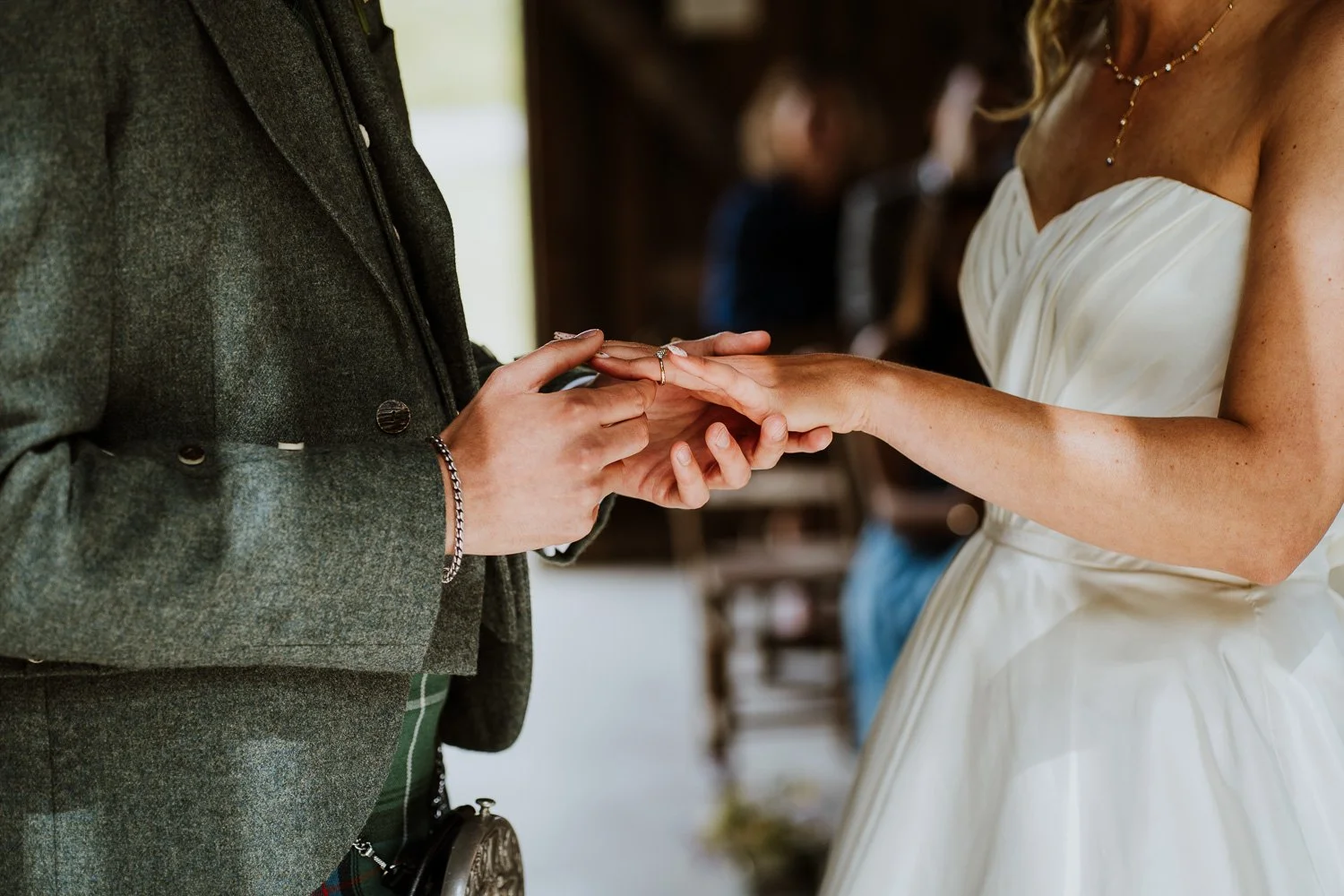 A couple exchanges rings during a wedding ceremony, with the groom in a gray jacket and the bride in a strapless white wedding dress, focusing on their hands.