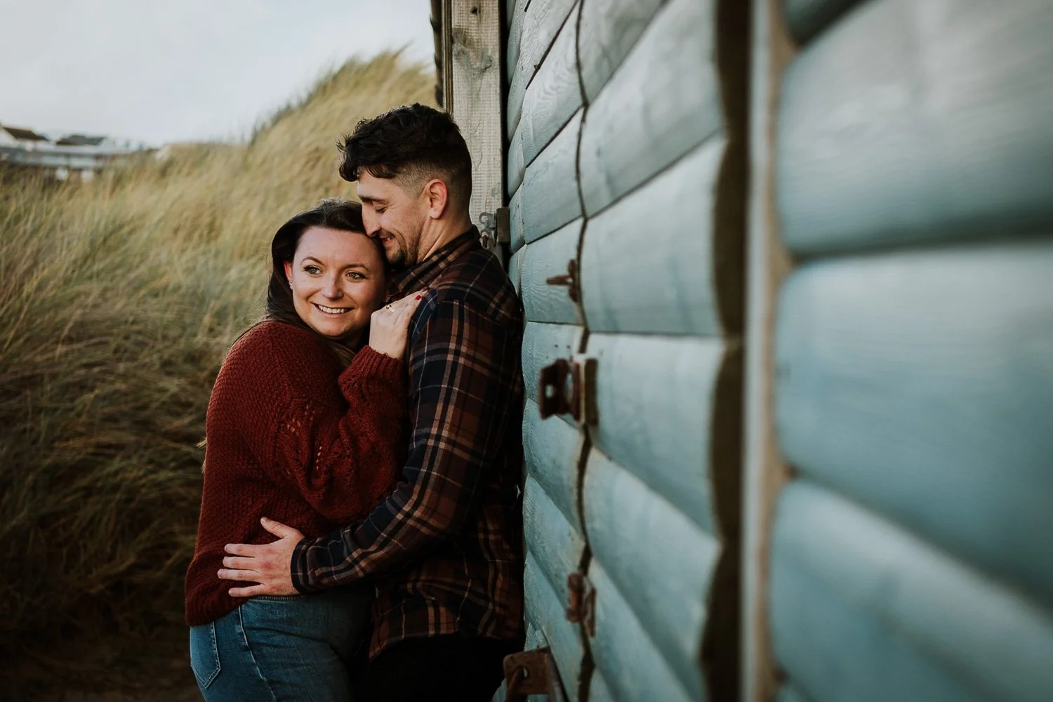 A happy couple hugging outdoors near a light blue wooden building, with sand dune grass in the background. Couple portrait taken during Engagement Photoshoot