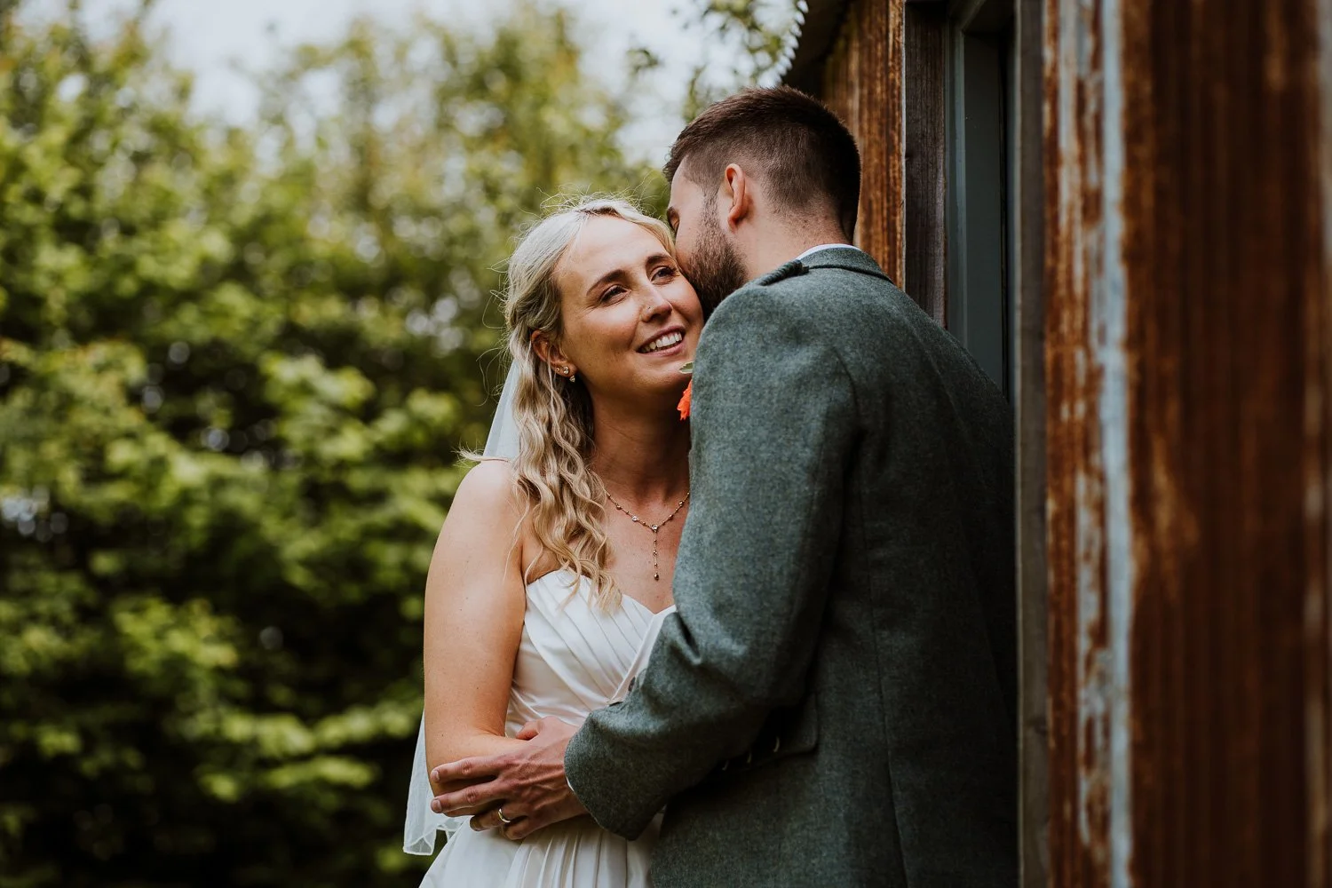 A bride and groom standing outdoors near a wooden building, sharing an intimate moment, with the bride smiling and the groom leaning in.