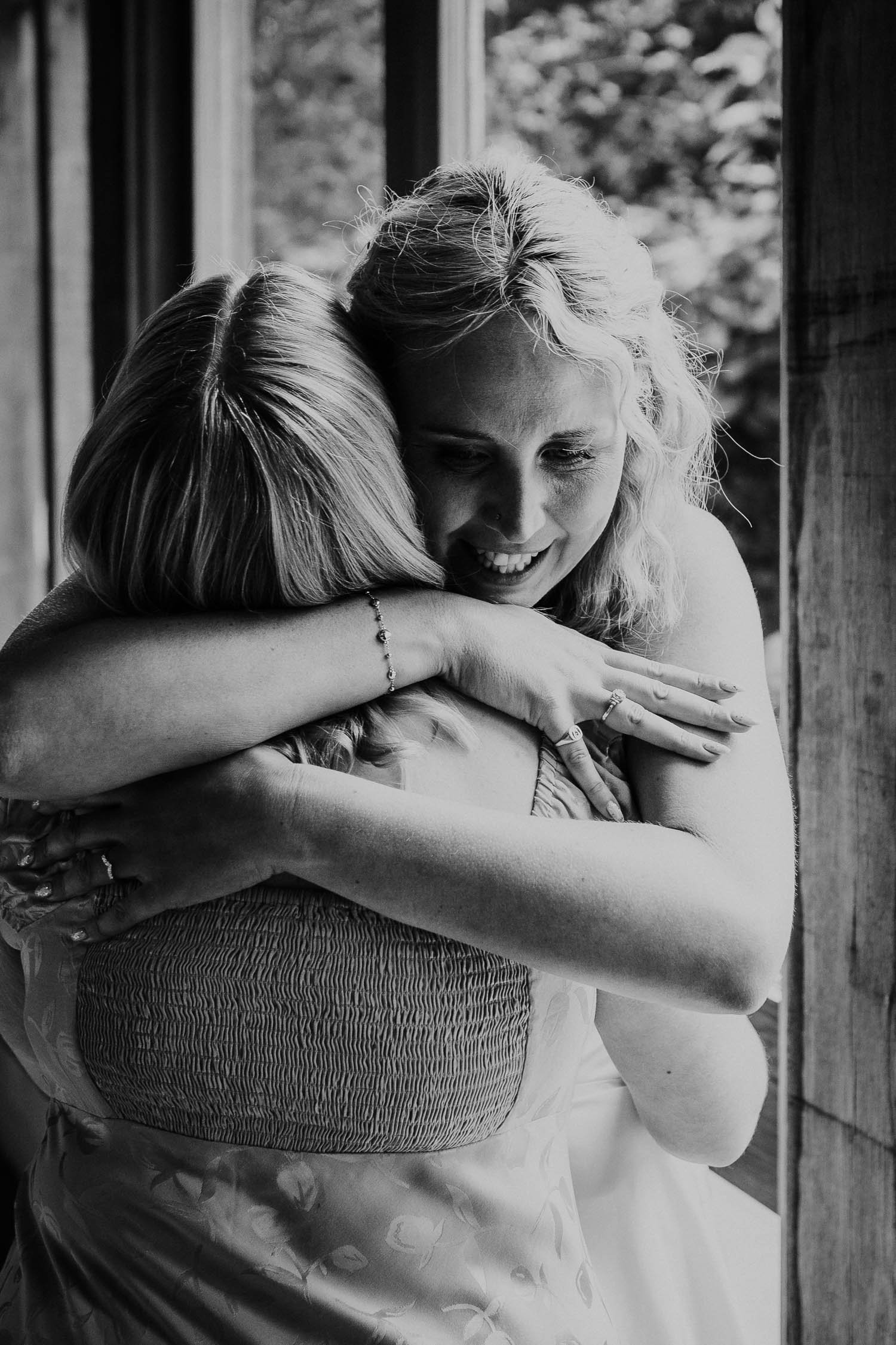 Two women hugging tightly inside a room, with a window and outdoor trees visible in the background. One woman, with curly hair, is smiling warmly.
