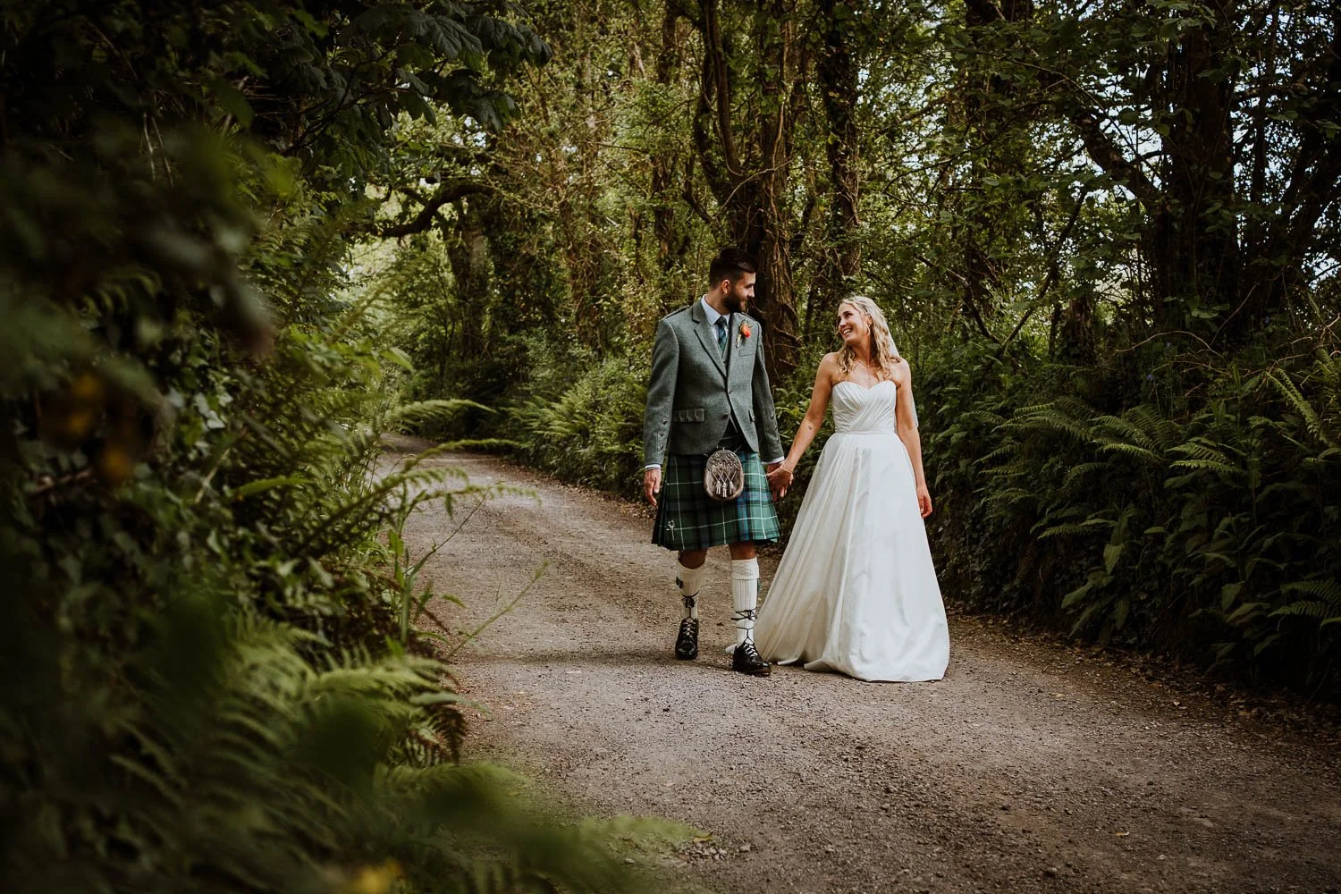 A bride and groom walking hand in hand on a forest trail, smiling at each other, surrounded by lush greenery. Natural Wedding Couple Portrait
