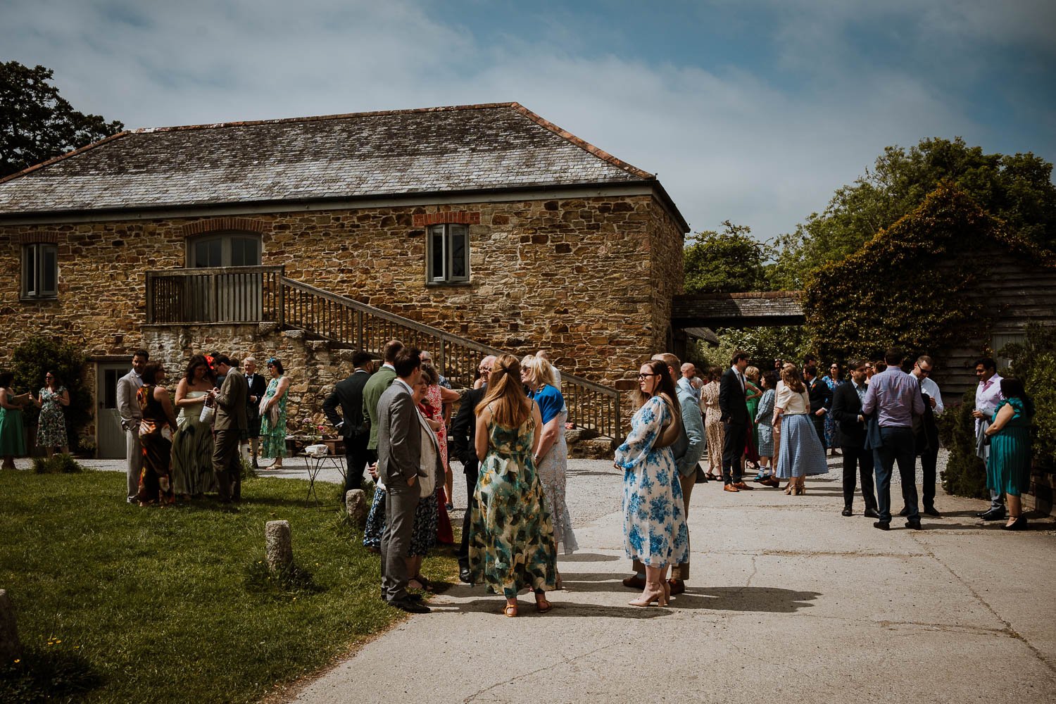 Wedding guests gathered outdoors in front of a rustic stone building on a sunny day. Wedding guests arrival