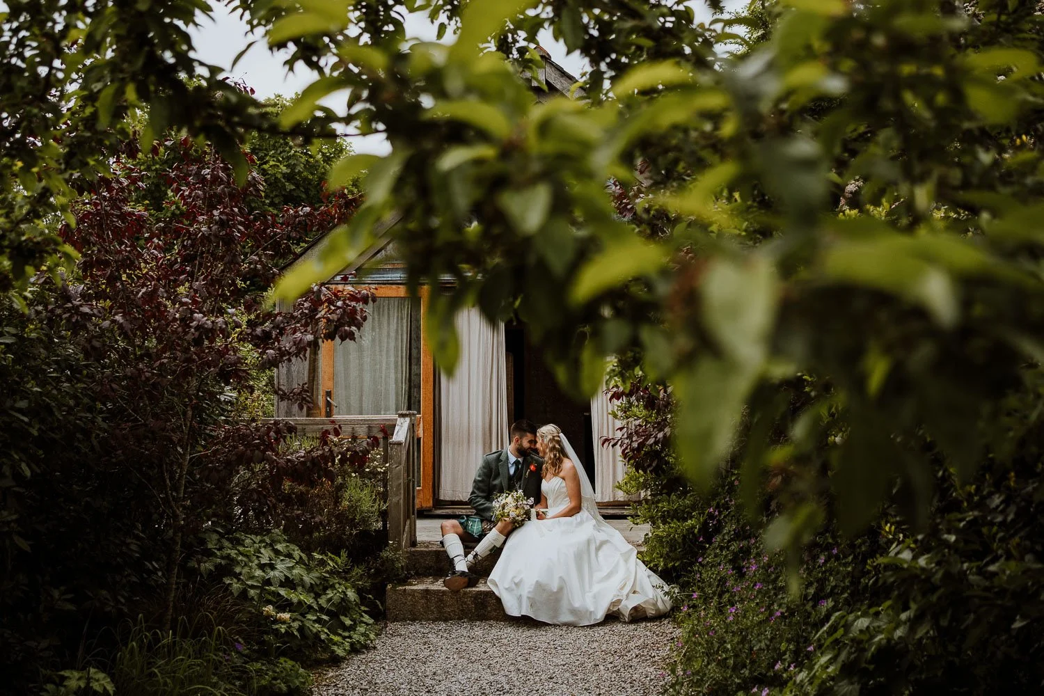 A bride and groom sitting on steps of their accommodation for their wedding day , sharing an intimate moment, surrounded by lush greenery.