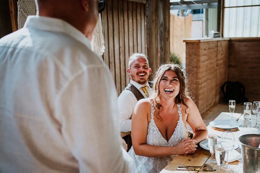 Bride and groom laughing at a wedding reception, father of the bride in foreground during his speech