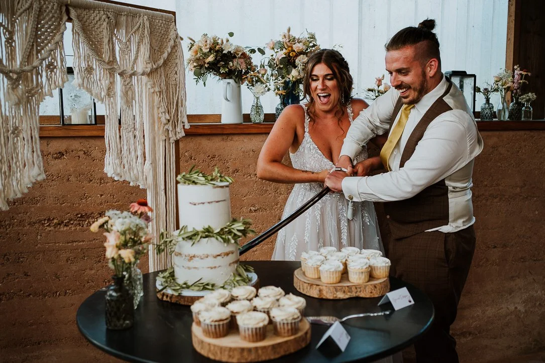Bride and groom cutting a wedding cake together, with cupcakes on the table, at their wedding reception.