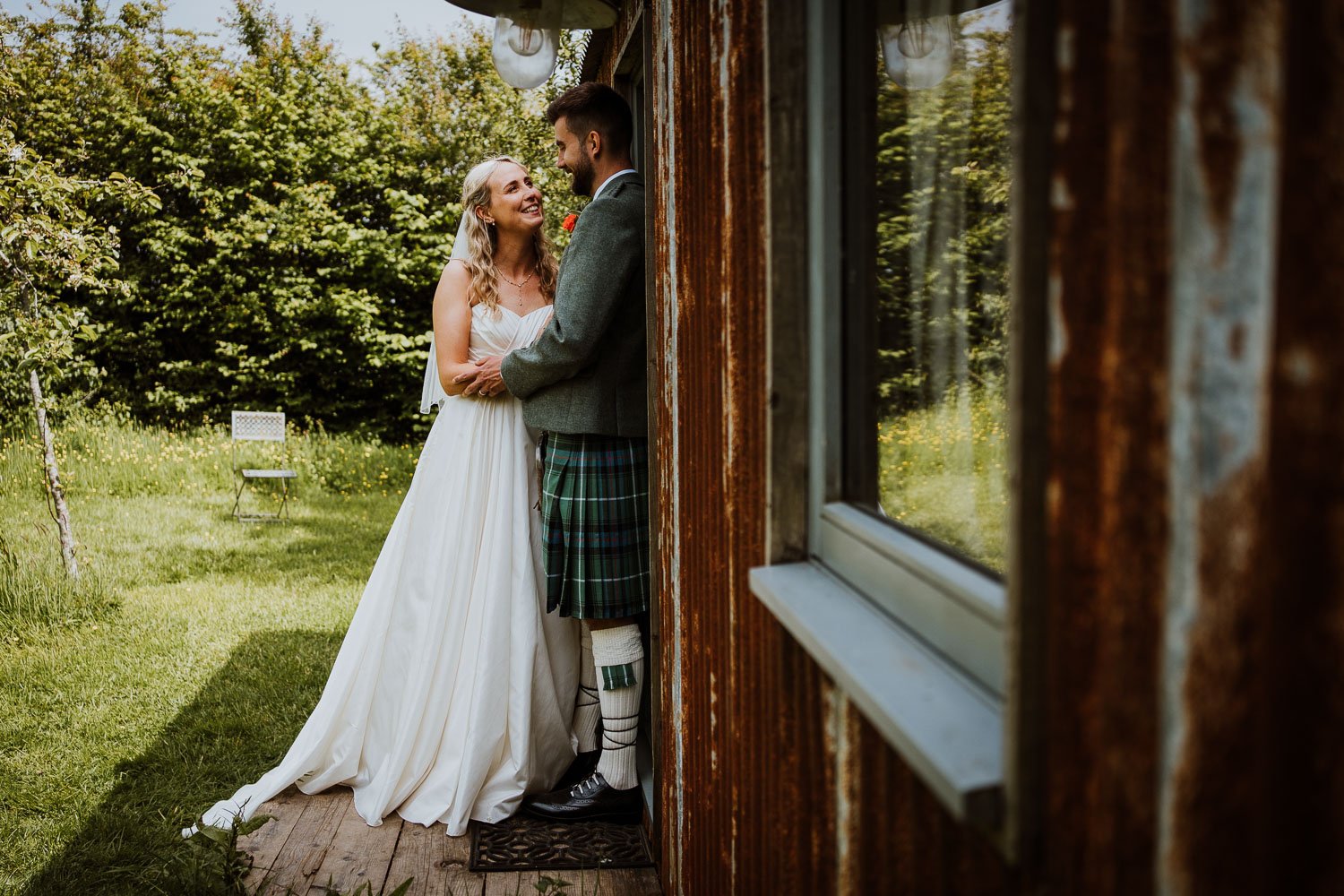 A bride and groom sharing a moment outside a rustic building on a sunny day. The bride is in a white wedding gown, and the groom is dressed in traditional Scottish attire, including a kilt and jacket. They are smiling and looking into each other's ey
