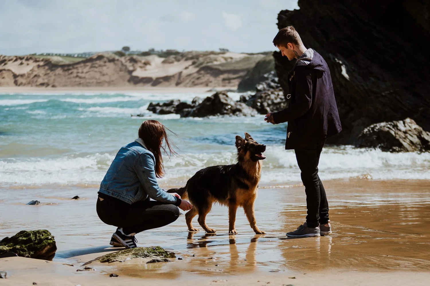 A couple with a German Shepherd dog on a beach with waves and rocky cliffs in the background. Natural Engagement Photos