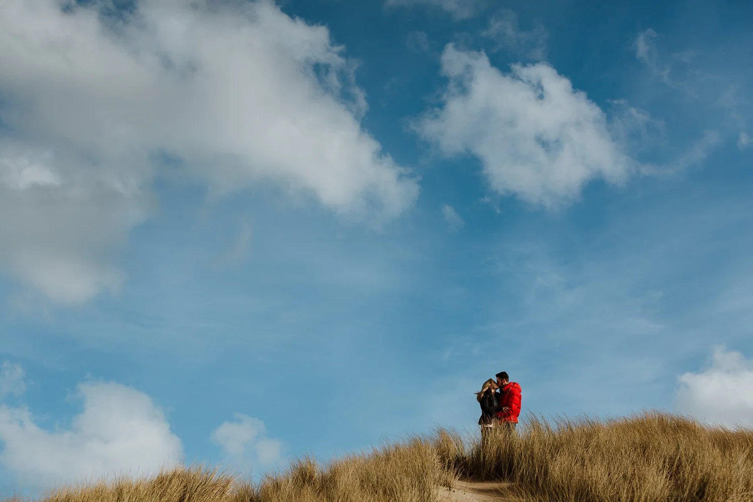 A couple standing on a sand dune, embracing and kissing under a partly cloudy blue sky. Engagement Photo. Couple Portrait