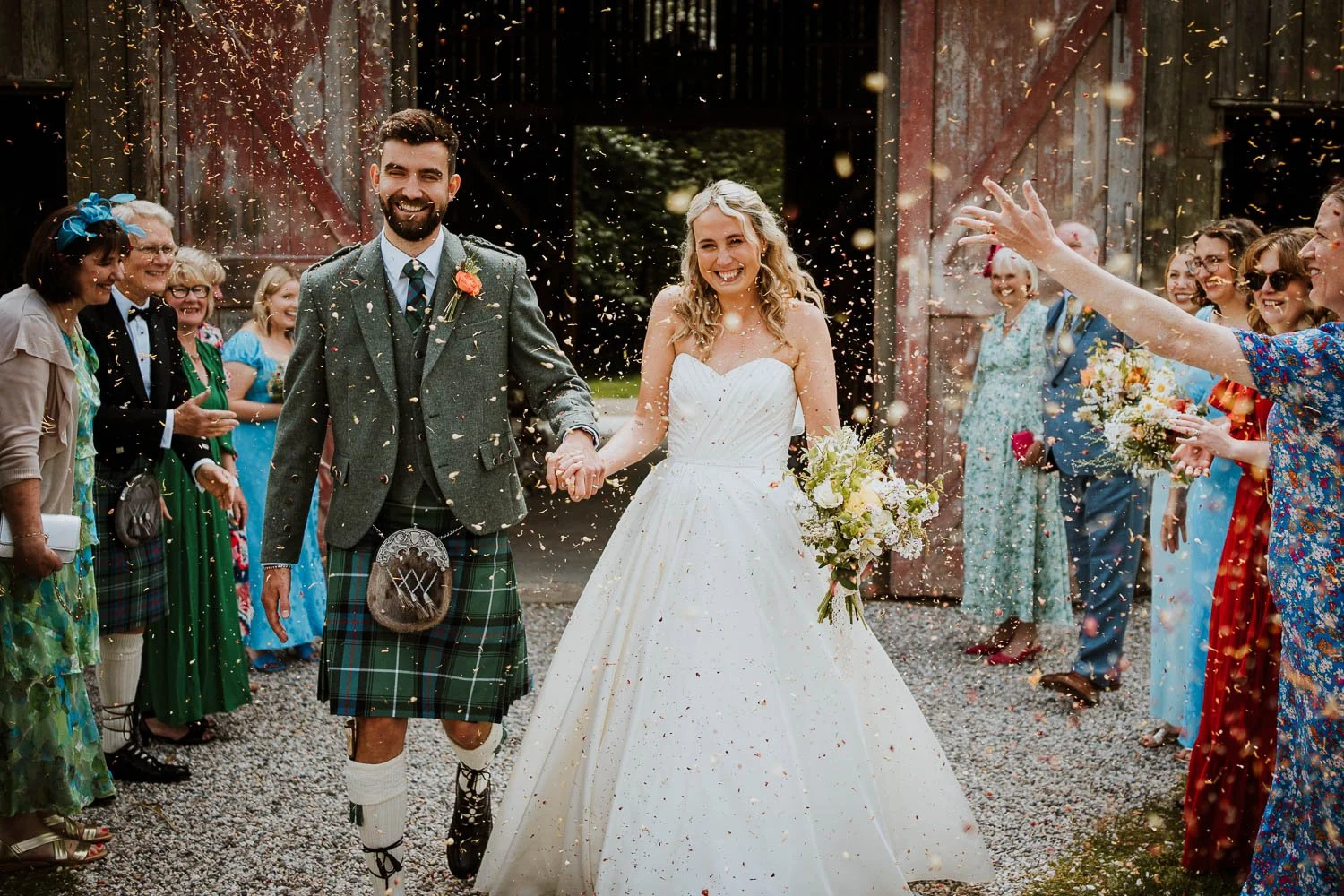 A bride and groom holding hands and smiling as they walk through a celebration crowd, with confetti falling around them outside the rustic barn at Nancarrow Farm