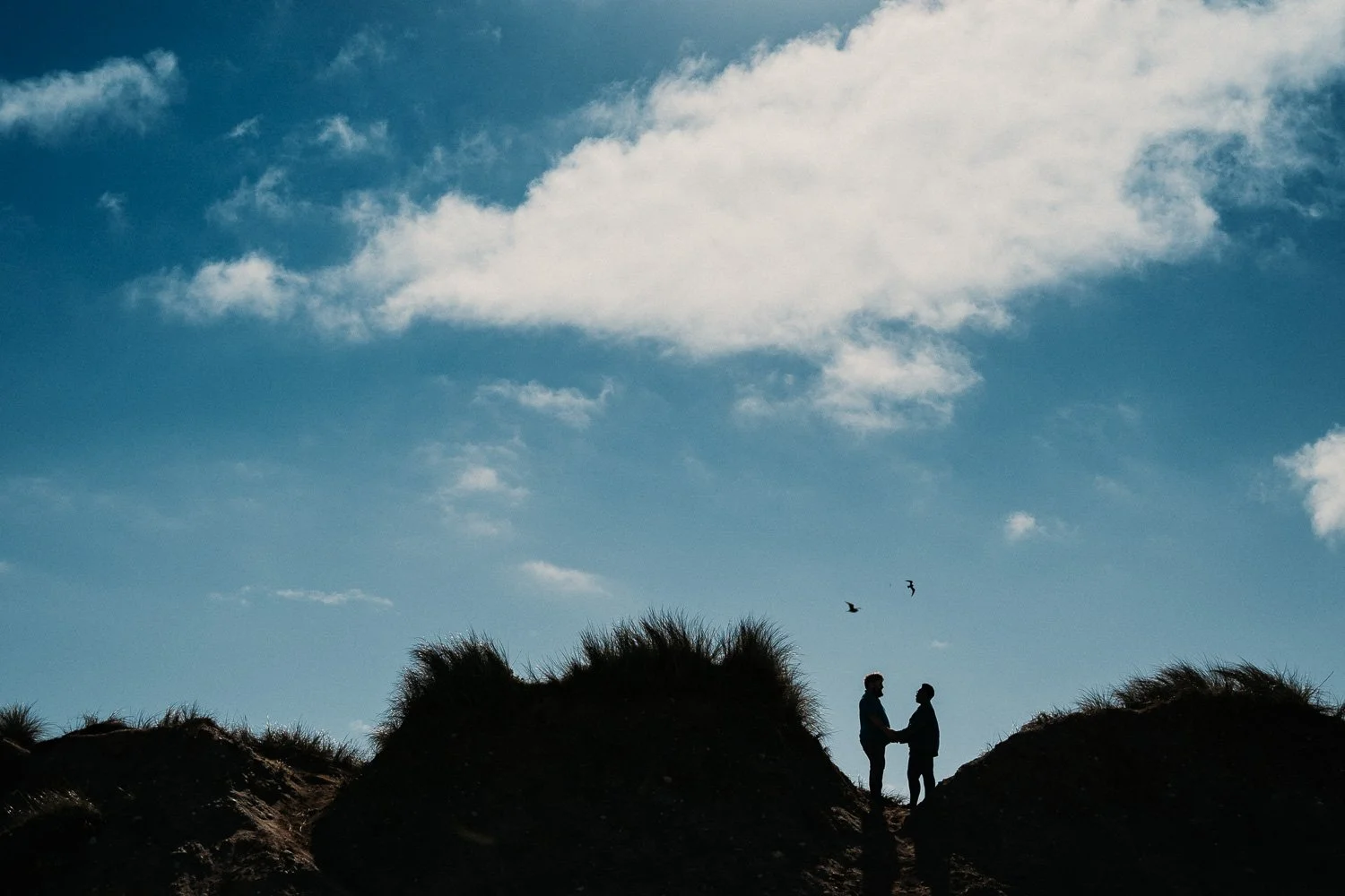 Silhouettes Portrait of a couple on a sand  dune at sunset, with a blue sky, clouds, and birds flying overhead. Couple portrait taken during Engagement Photoshoot