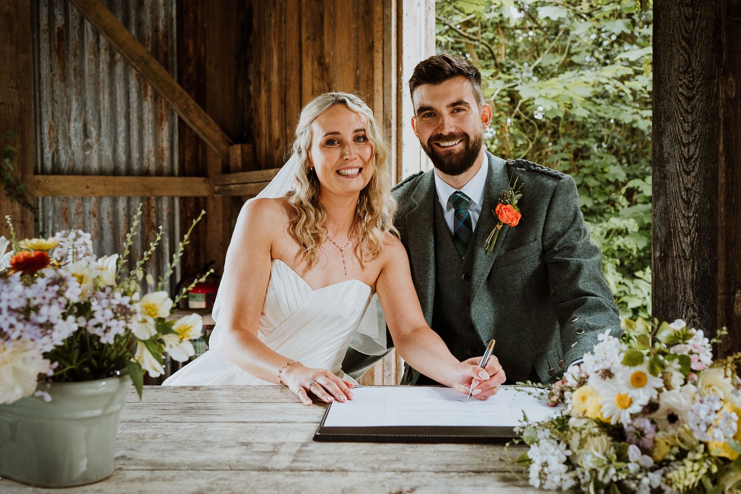 A bride and groom signing their wedding registry or marriage license at a rustic wooden table, surrounded by flowers, inside a barn with natural light and greenery in the background.