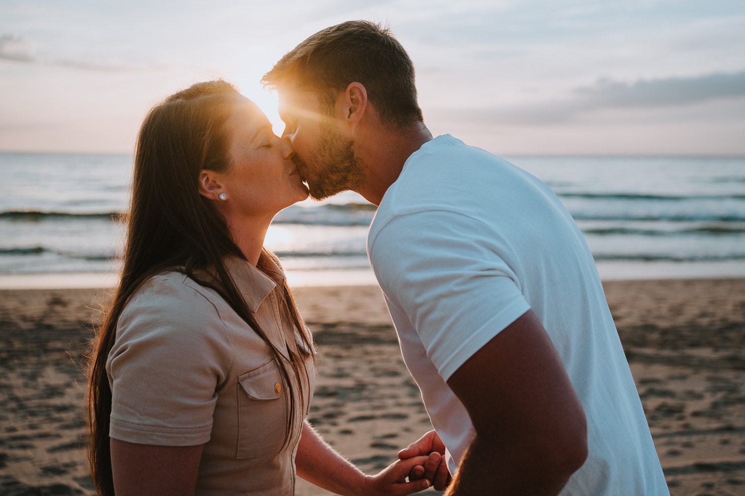 A couple kissing on the beach at sunset, holding hands with the ocean and sky in the background. Couple portrait taken during an engagement photo session