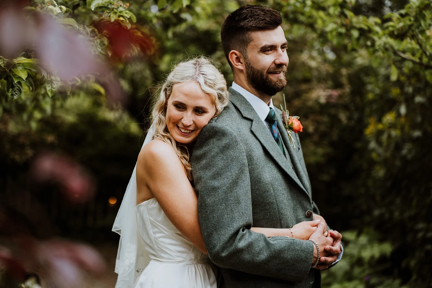 A bride and groom stand close together outdoors, the bride hugging the groom from behind with a smile, surrounded by greenery and flowers. Natural Wedding Couple Portrait