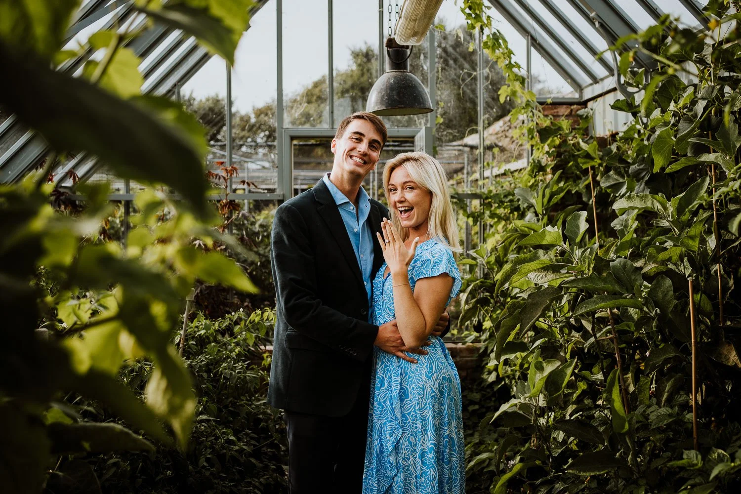 A joyful couple celebrating an engagement in a greenhouse filled with lush green plants, with the woman showing off her engagement ring. Natural Proposal Photography