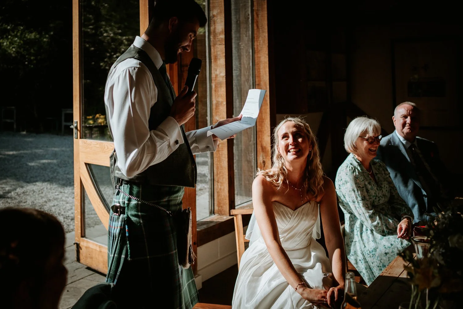 A man in a kilt giving a speech at a wedding reception, with the bride and seated guests smiling and enjoying the moment.
