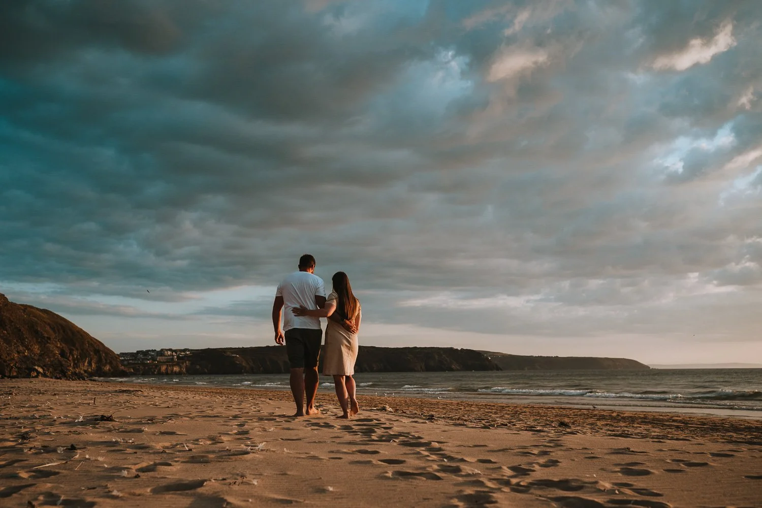 Couple walking along the beach at sunset with stormy clouds overhead. Couple portrait taken during an engagement photo session