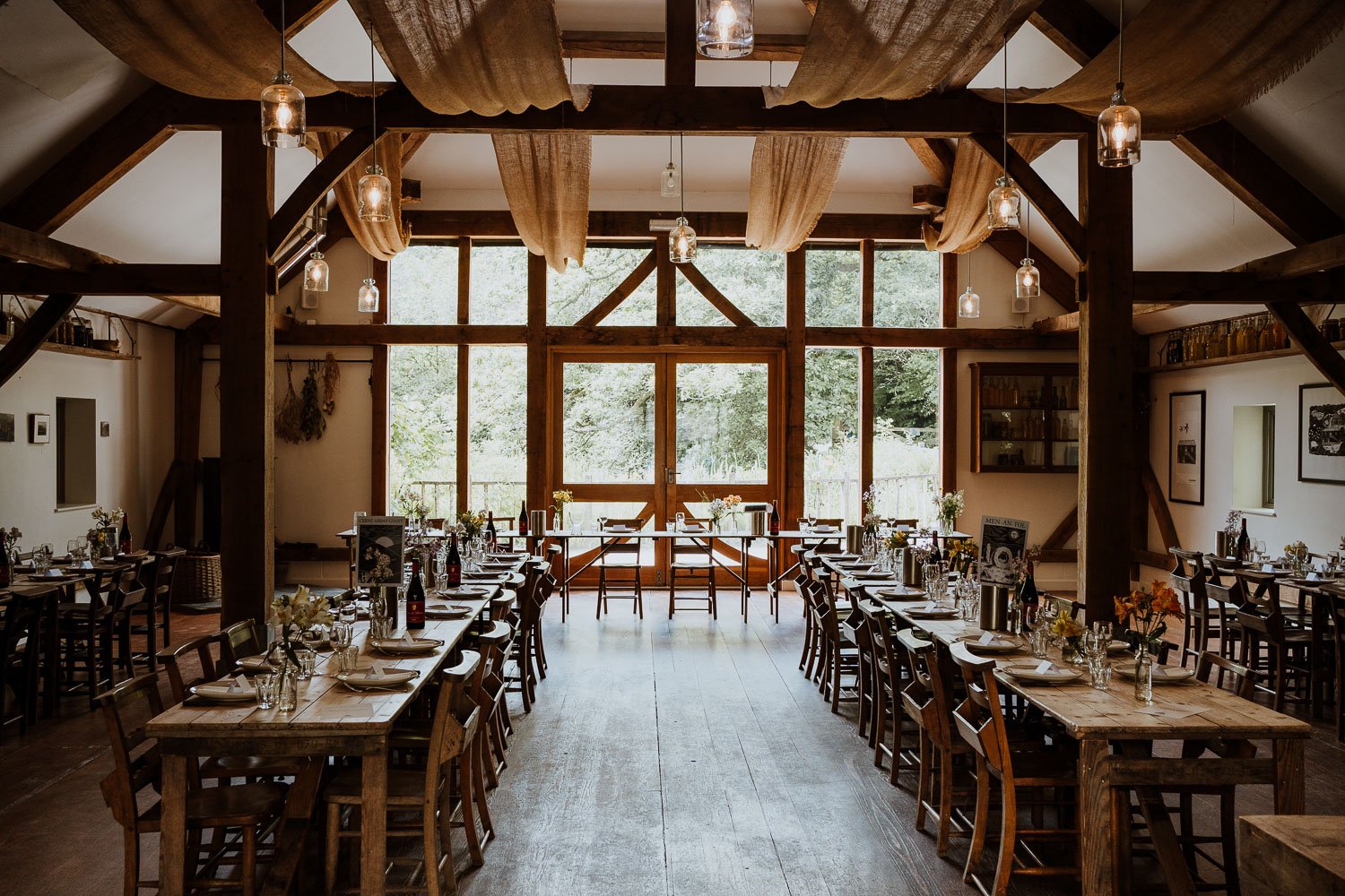Interior of a rustic wedding reception barn with wooden tables, chairs, and windowed doors leading outside, decorated with flowers and candles.