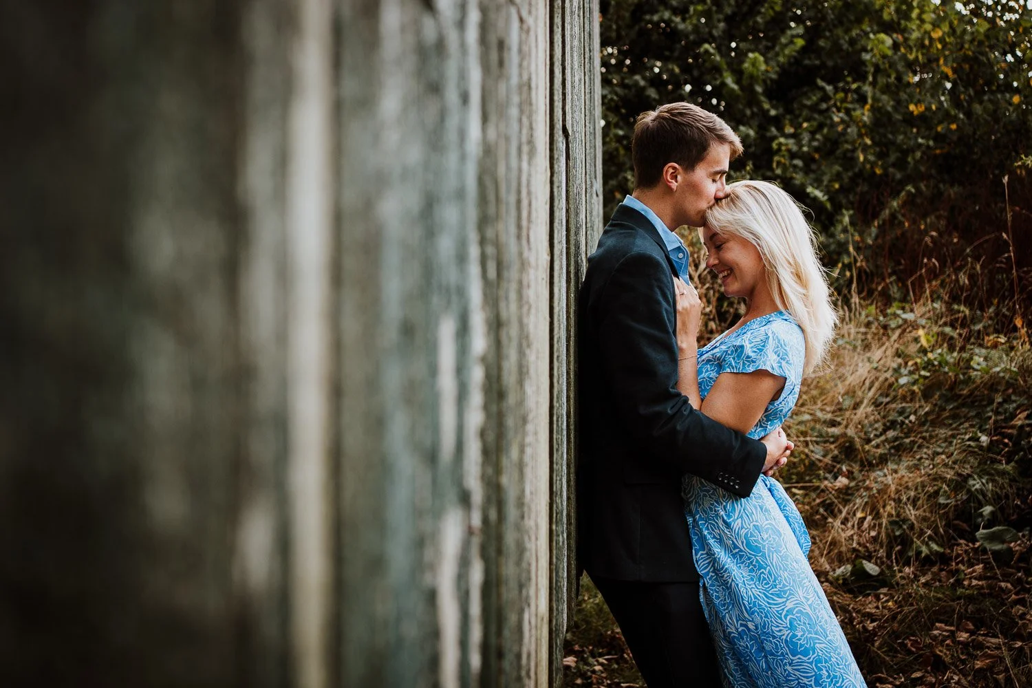 A couple sharing a romantic moment outdoors, standing close against a weathered wooden wall, with trees and foliage in the background. Proposal Photography, Natural Couple Portrait