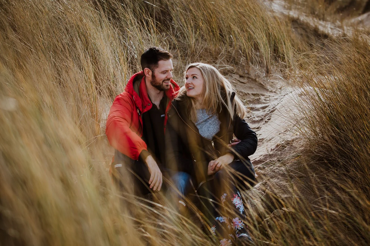 A couple sitting on a sandy dune surrounded by tall grass, smiling at each other during sunset. Couple Portrait taken during Engagement Photo Session