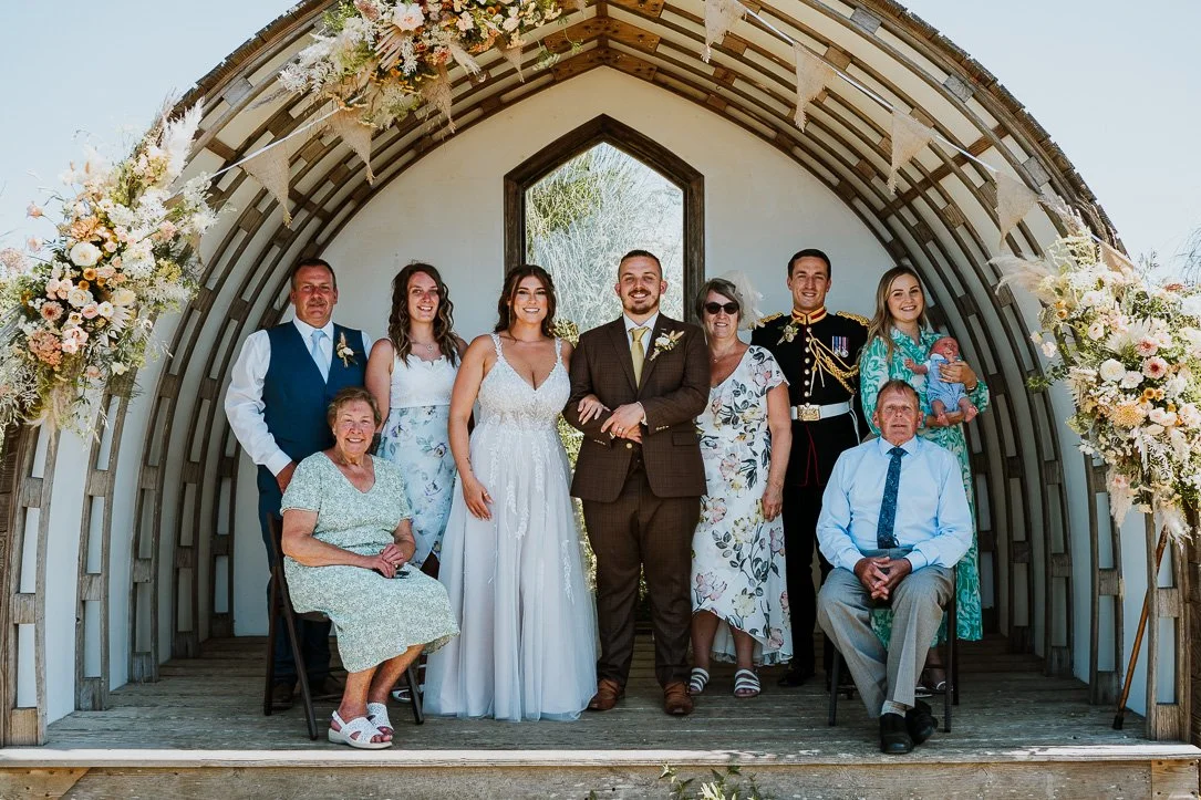 A wedding group photo with nine people, including the bride in a white dress, the groom in a brown suit, and others in formal attire, under a decorated wooden arch with flowers.