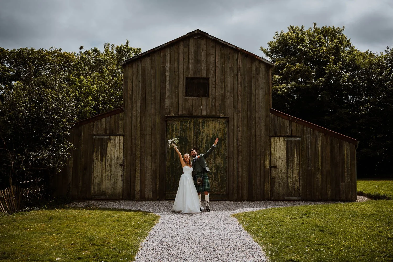 A bride and groom celebrating in front of a rustic wooden barn, with the bride holding a bouquet and both raising their arms in joy.