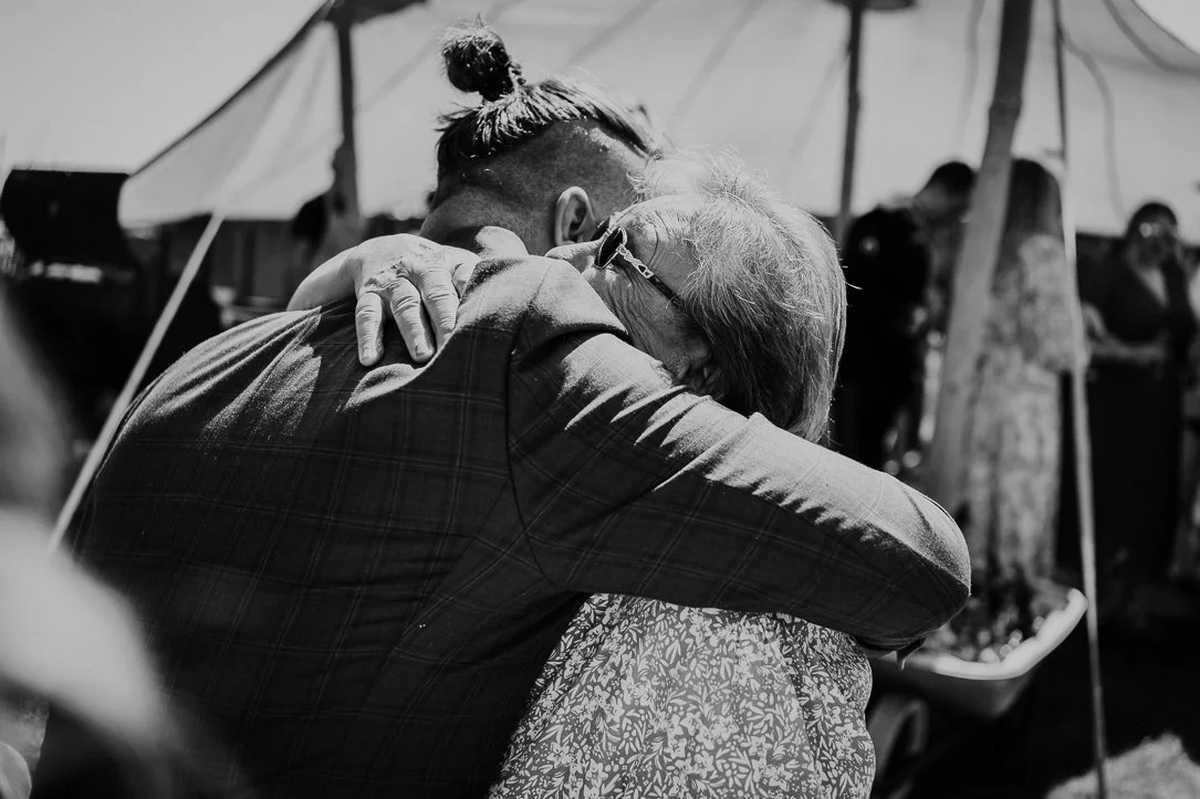 Two people hugging in an outdoor setting, with a tent and trees in the background.