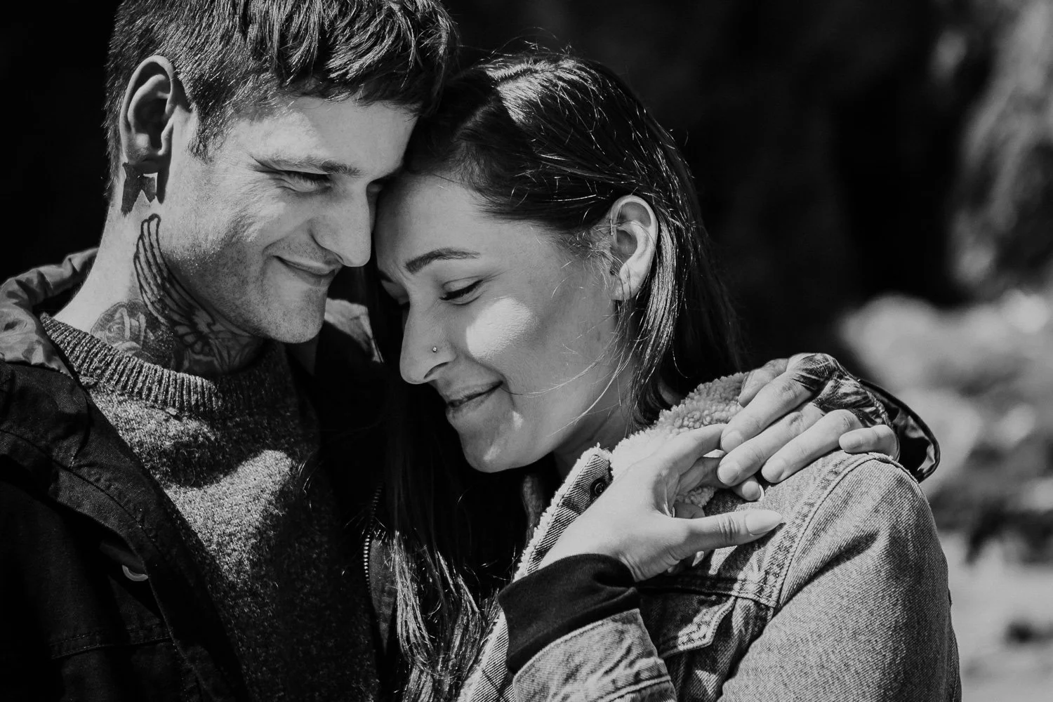 A black and white close-up photo of a young couple enjoying a tender moment outdoors, with their foreheads touching and eyes closed, smiling softly. Natural Engagement Photos