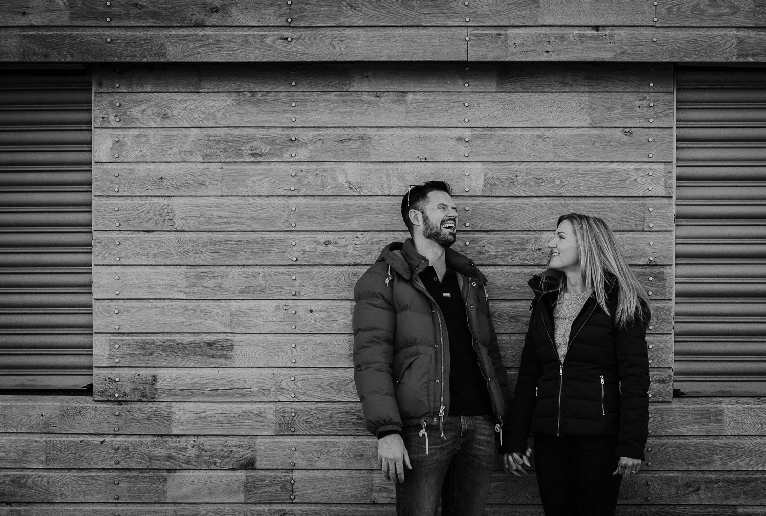 A man and a woman standing outdoors in front of a rustic wooden wall, holding hands and smiling at each other. Couple Portrait taken during Engagement Photo Session