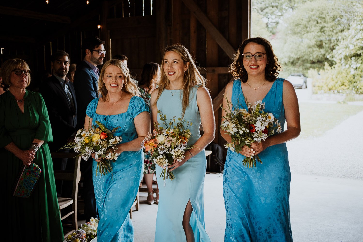 Three women in blue dresses holding bouquets walking in a wedding ceremony, with other guests in the background inside a rustic barn.