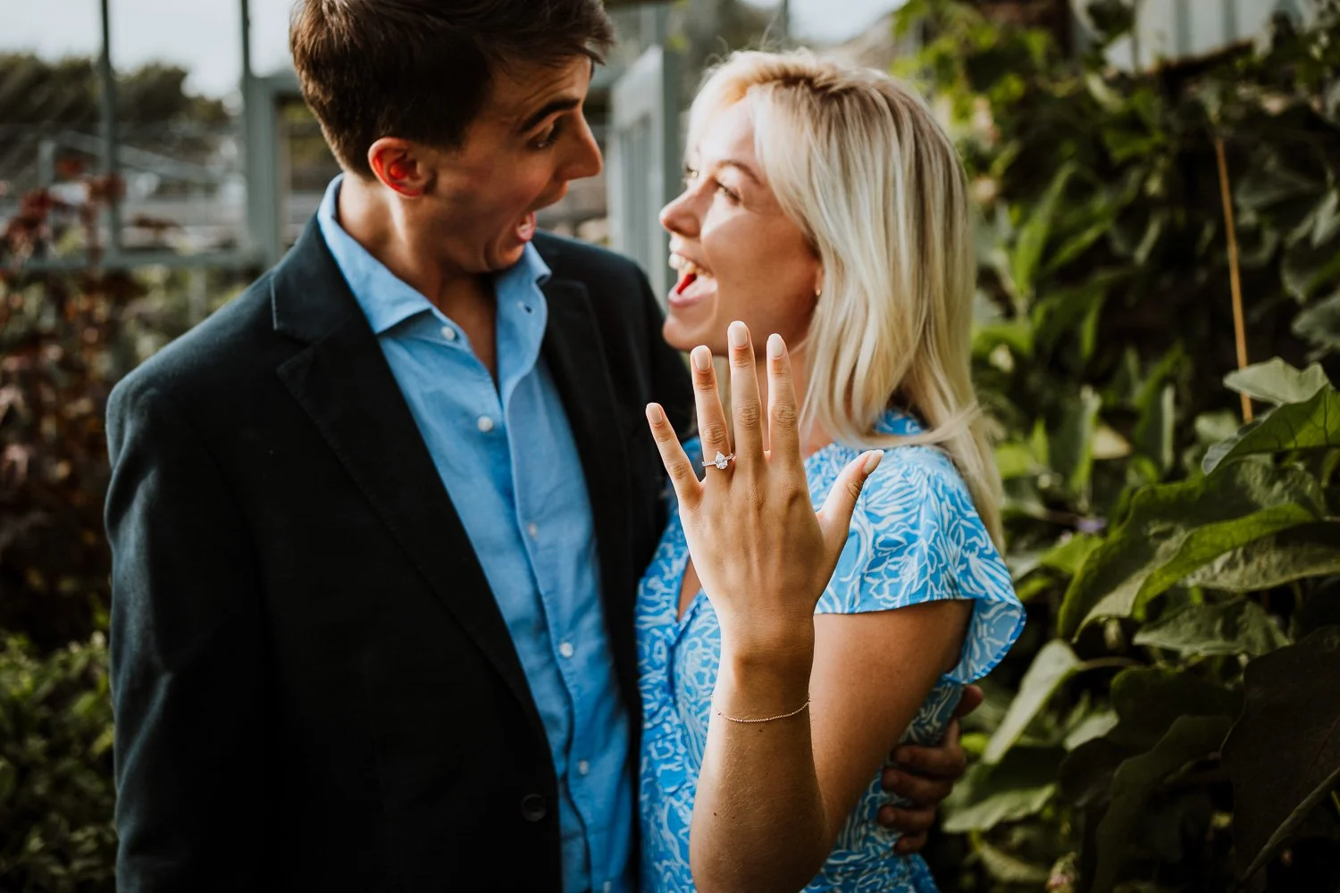 A man and woman are celebrating an engagement, with the woman showing her ring and both smiling, standing among greenery. Natural Proposal Photography