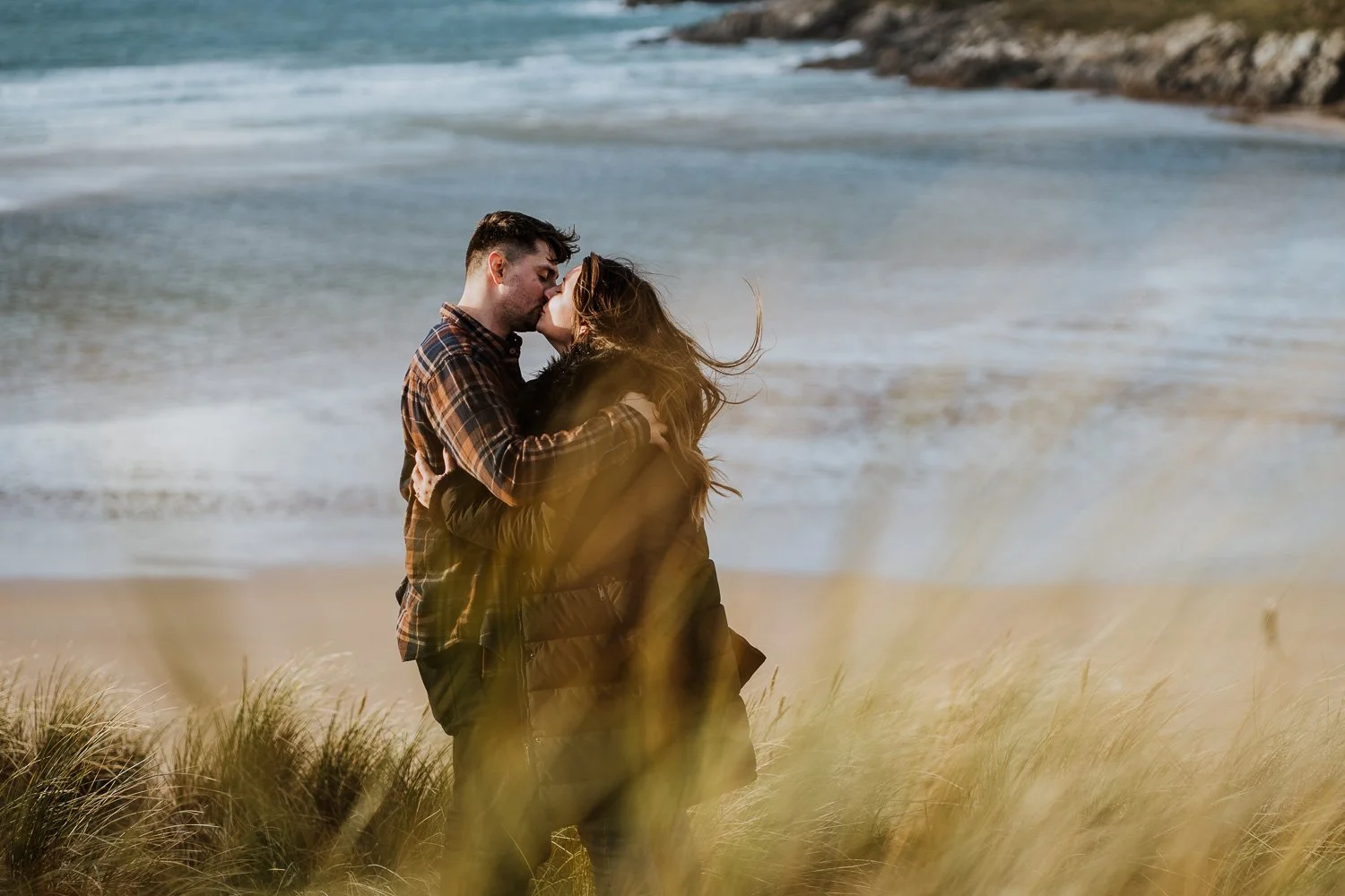 A couple embraces on a sandy beach with tall grasses, sharing a kiss near the shoreline with waves in the background. Engagement Photo of couple on the beach in Cornwall