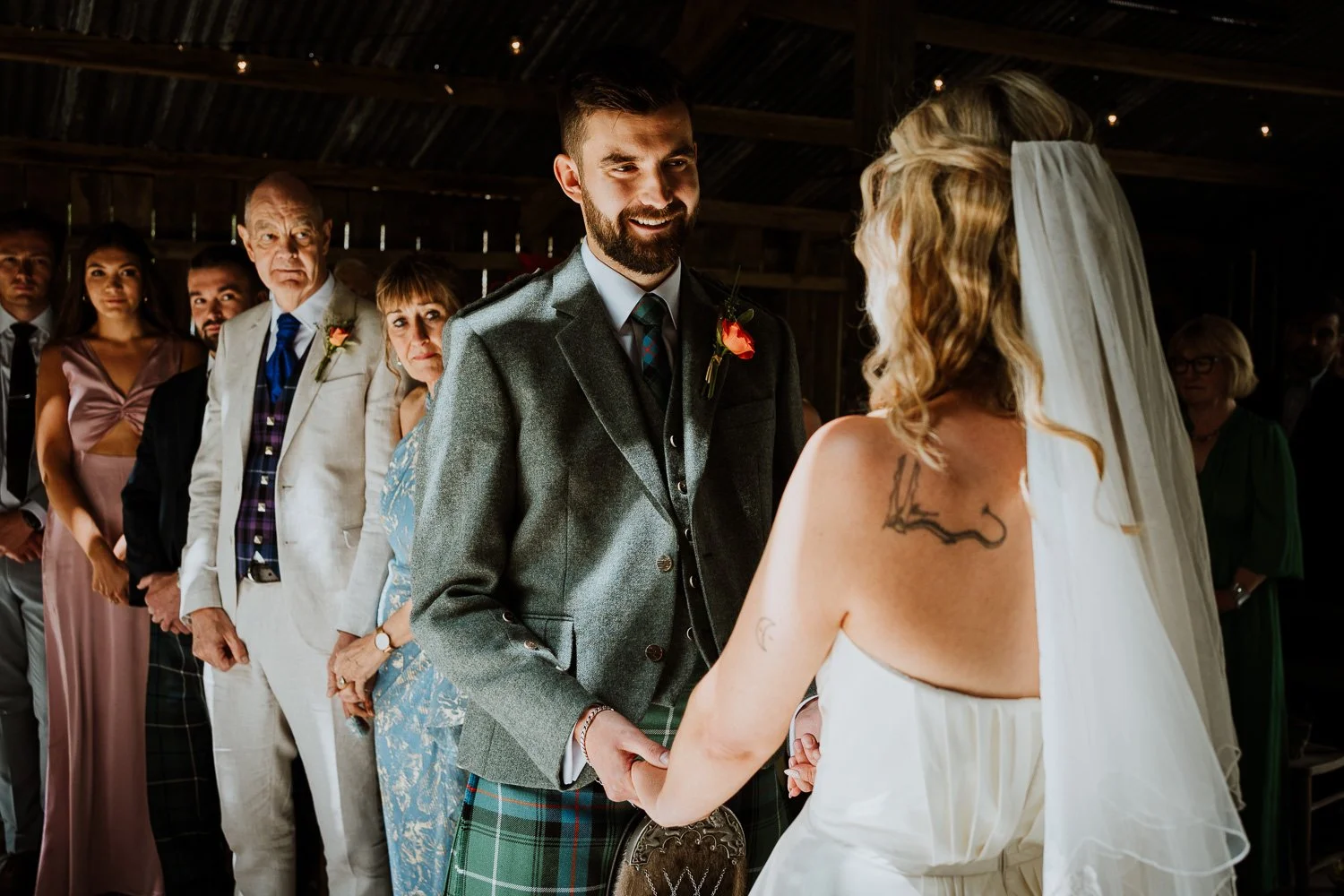 A wedding ceremony with a groom in a gray suit and a bride with tattoos, wearing a white dress and veil, holding hands. Guests are standing in the background.