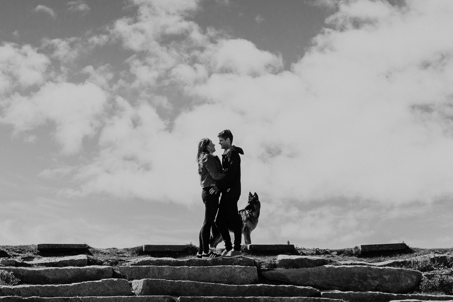 A black-and-white photo of a couple standing on stone steps on an outdoor hilltop, looking at each other and smiling. Their dog stands near them. The sky is partly cloudy. Natural Engagement Photos with dogs