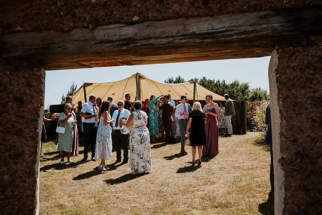 People gathered outdoors at a social event under a large beige canopy, seen through a stone or brick archway.