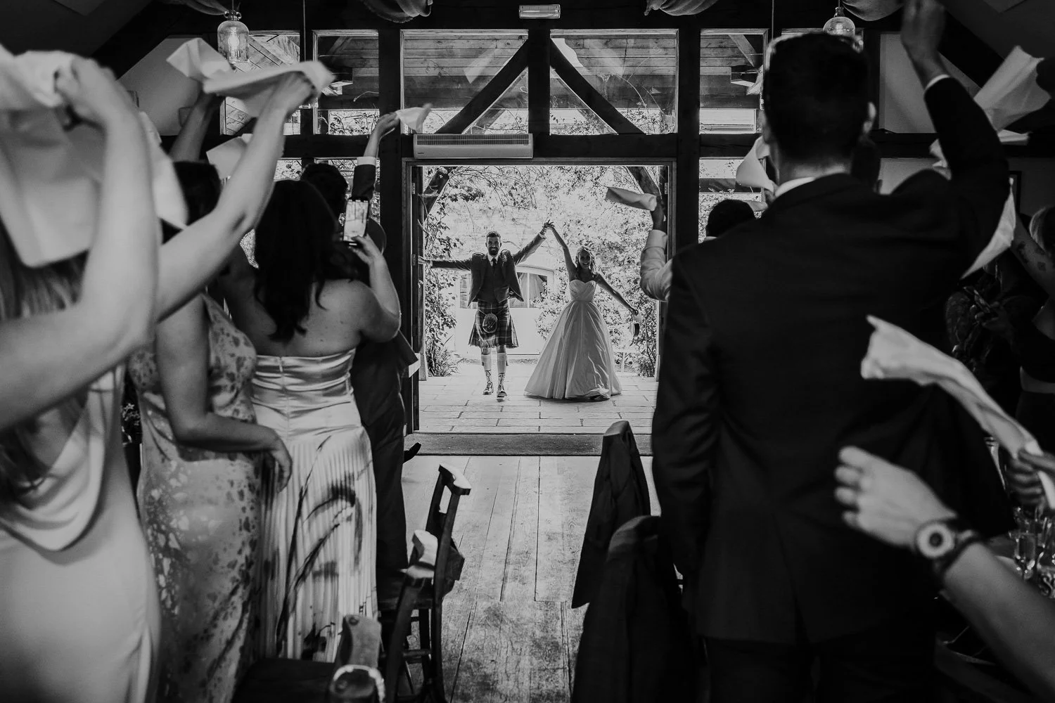 Bride and groom holding hands and raising arms as they walk out of a venue, with guests clapping and taking photos inside, black and white photo.