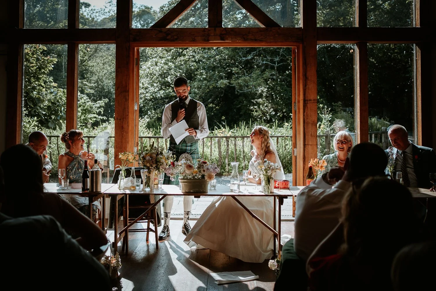 A wedding reception inside a wooden venue with large windows. The bride and groom are sitting at a table, laughing as a man in traditional attire reads from a paper. Guests are seated around, some smiling and some with drinks, with sunlight coming in