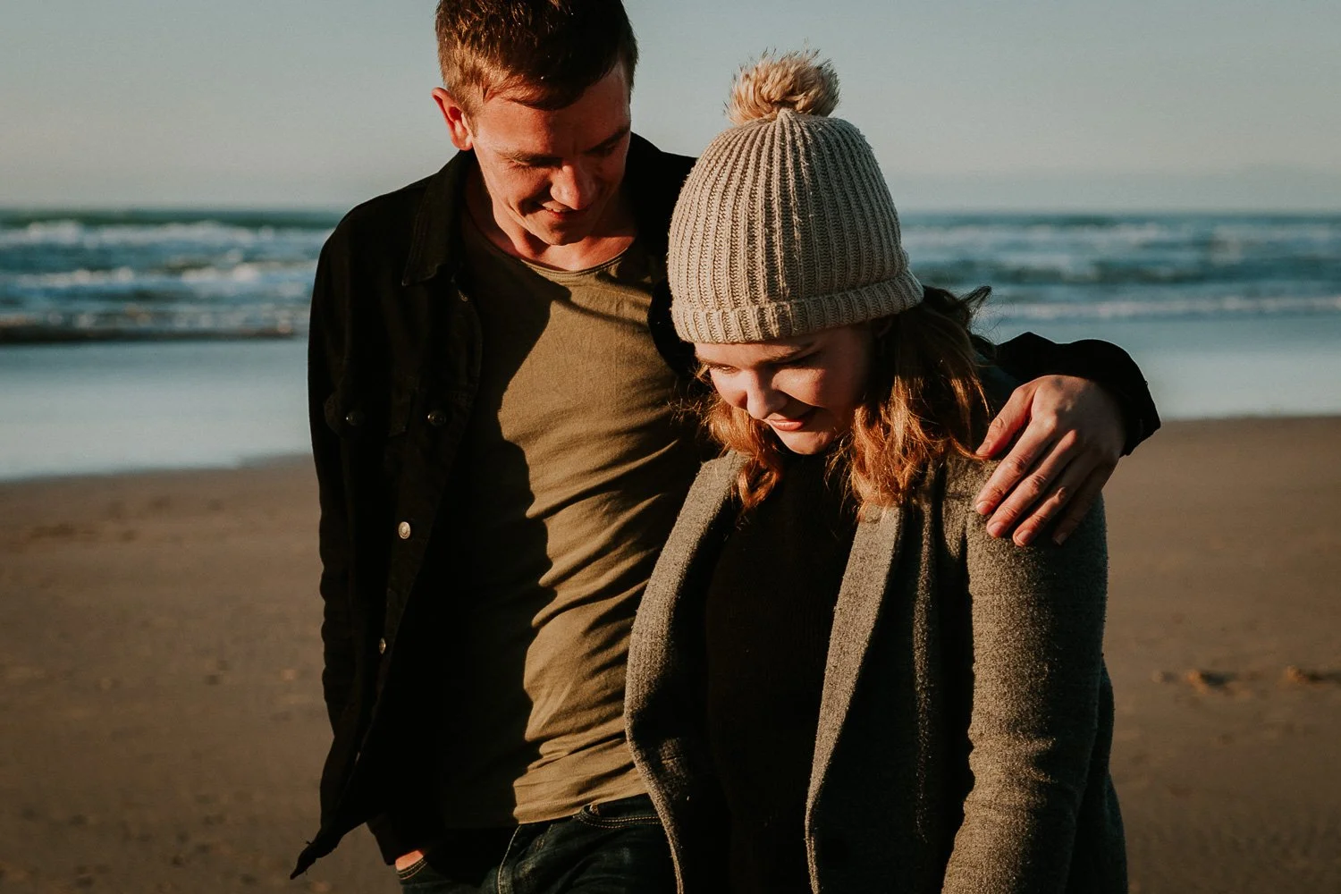 A young man and woman walk together on the beach at sunset, smiling and with the man's arm around the woman's shoulder. Couple portrait taken during an engagement photo session