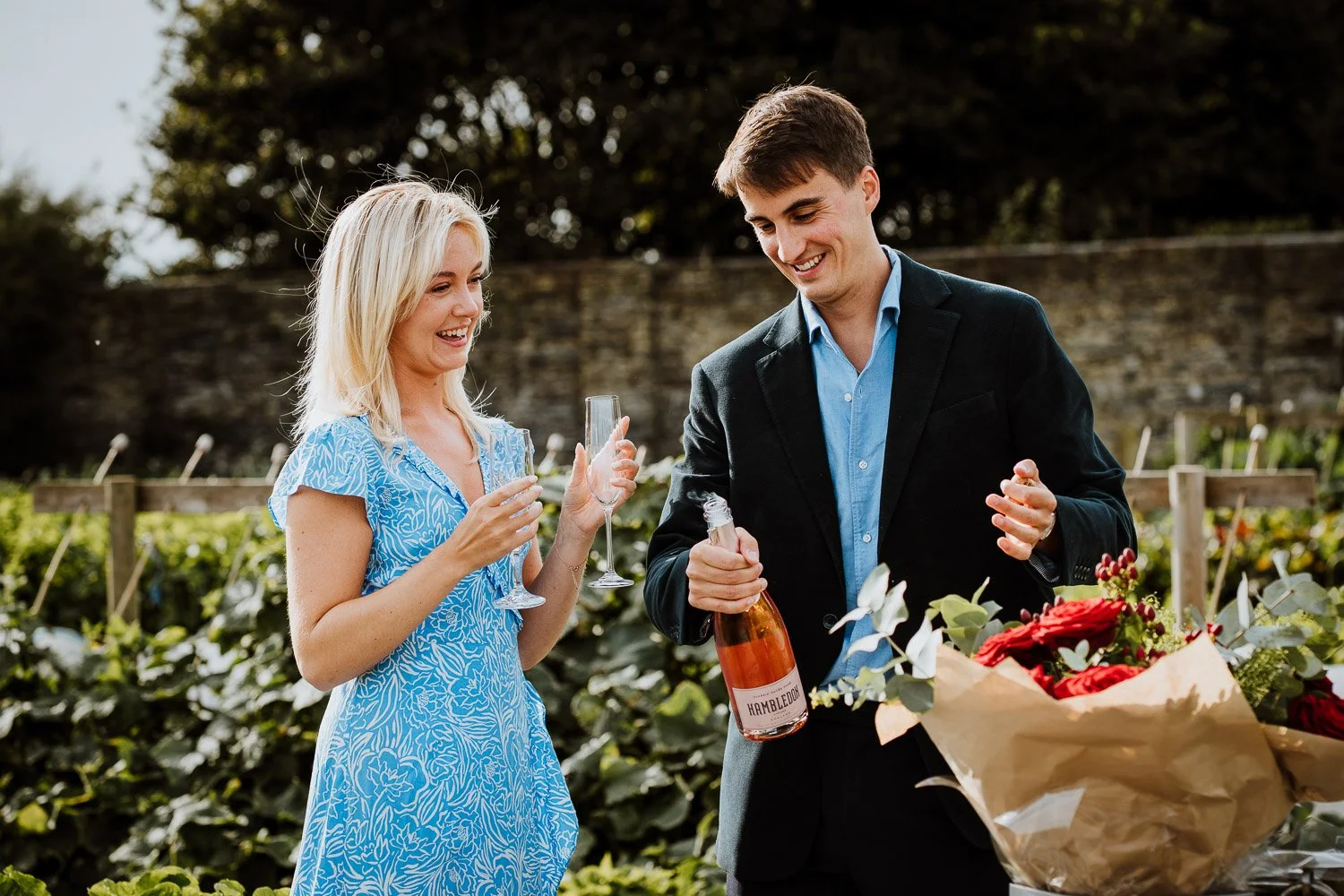 A woman and a man smiling outdoors, woman holding a champagne flute, man holding a bottle of rosé wine, with a bouquet of red roses in front of him. Natural Proposal Photography