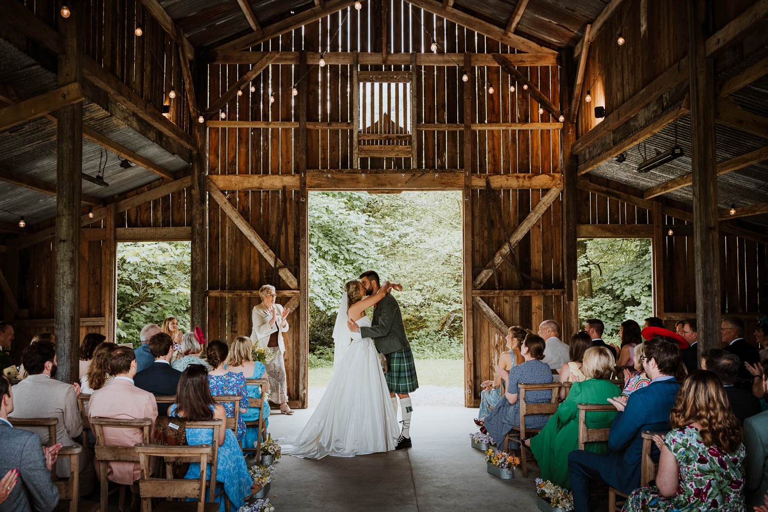 A wedding ceremony inside a rustic wooden barn with a bride and groom kissing at the altar, surrounded by seated guests and lush greenery outside.