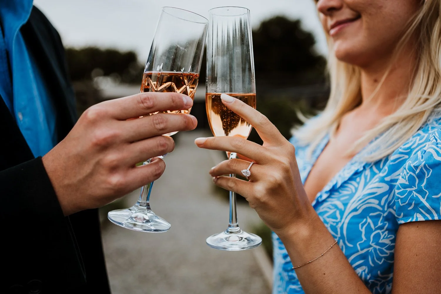 A couple toasting with glasses of rosé wine, with the woman wearing a ring on her finger and a blue patterned dress, celebrating outdoors. Proposal Photography