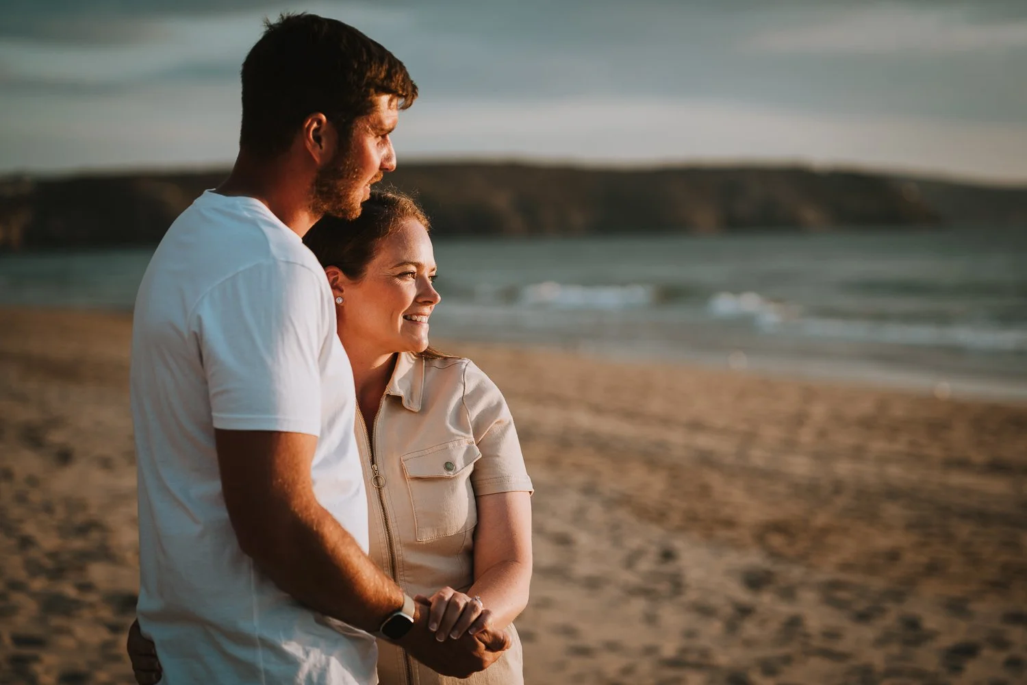A smiling couple standing on the beach during sunset, facing right with waves and hills in the background. Couple portrait taken during an engagement photo session