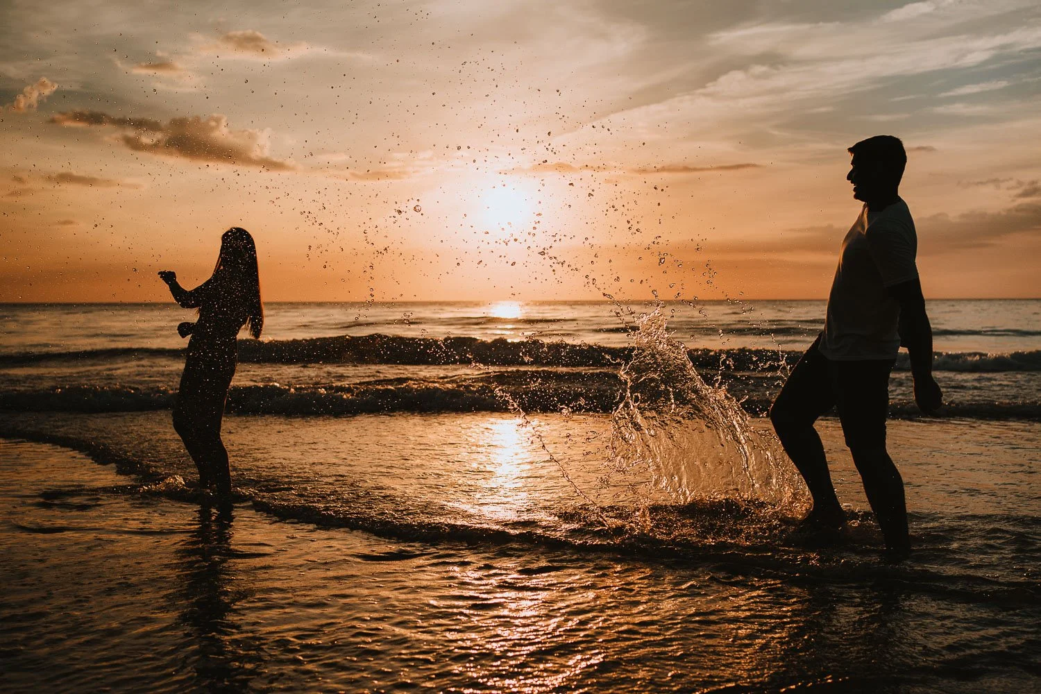 A silhouette of a man and a woman playing in the ocean near the shore at sunset, with water splashing at each other.  Playful, natural couple portraits taken during pre wedding photo / engagement photo session