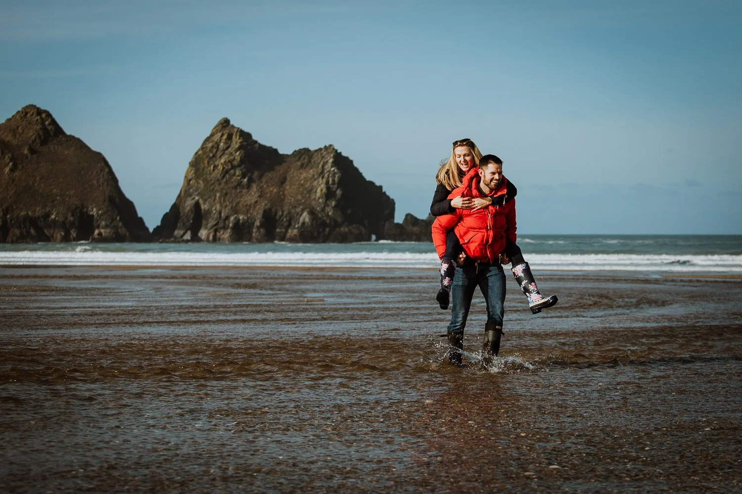 A couple enjoying a playful moment on a beach, with the man carrying the woman on his back, both smiling, during a clear day with rock formations in the background. Natural and Fun Couple Portrait taken during Engagement Photo Session
