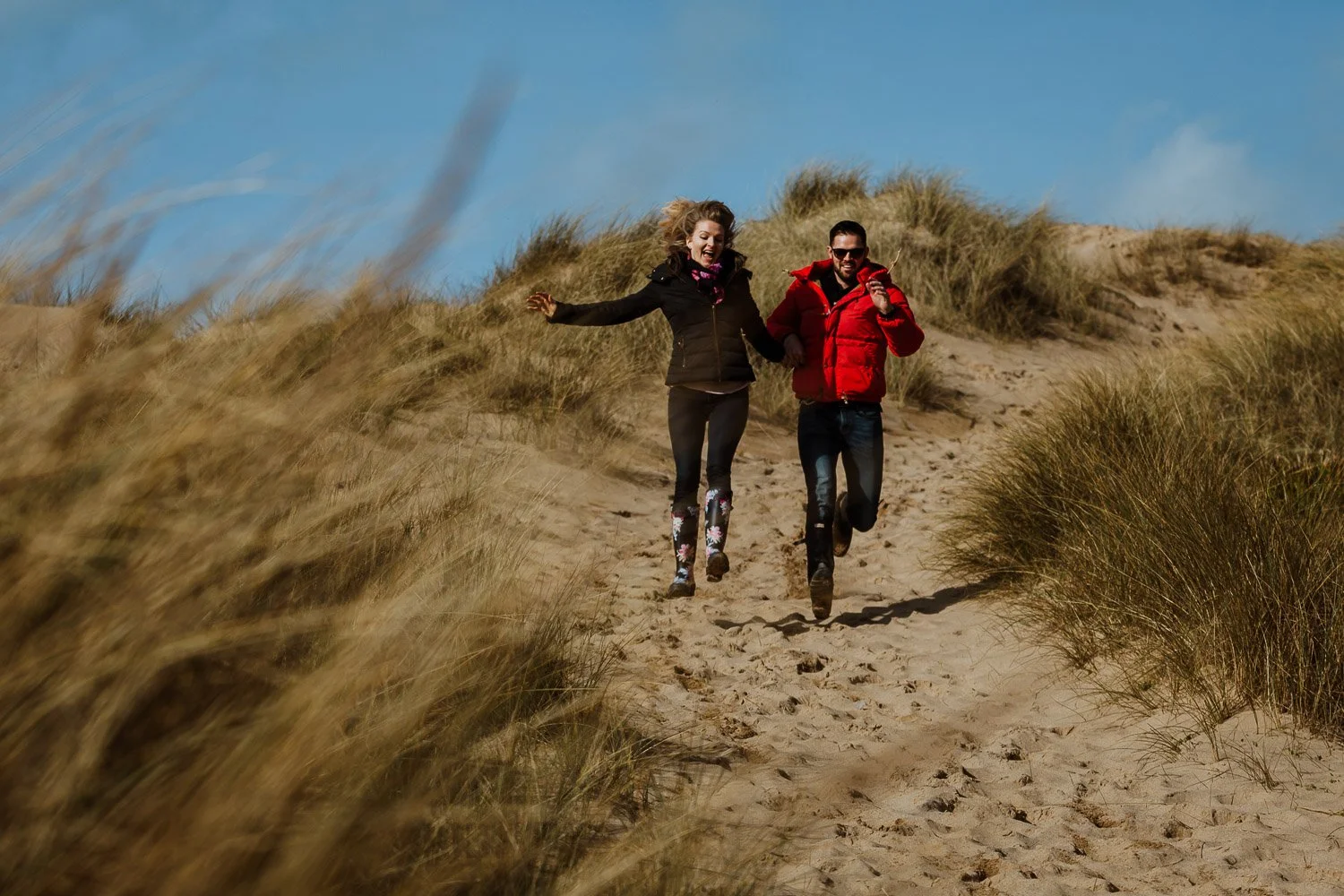 A couple running down sandy dunes with grass on a windy day. Natural, fun Couple Portrait taken during Engagement Photo Session