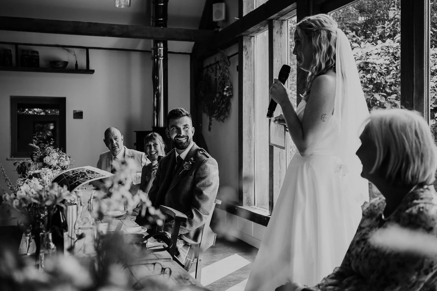 A bride giving a speech at her wedding reception, standing next to a table with guests including an older man, a woman, and a smiling man in a suit, in a rustic venue with wooden beams and large windows.