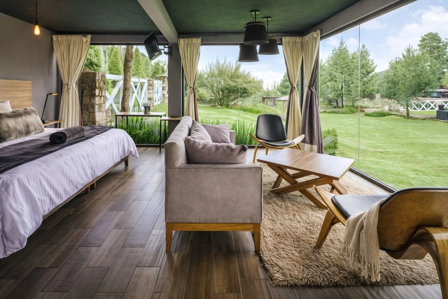 Interior de una habitación con grandes ventanas que muestran un paisaje montañoso con árboles y cielo azul. Hay un sofá negro de cuero, una mesa de madera y una cama con cojines y un tapete de felpa en el suelo.