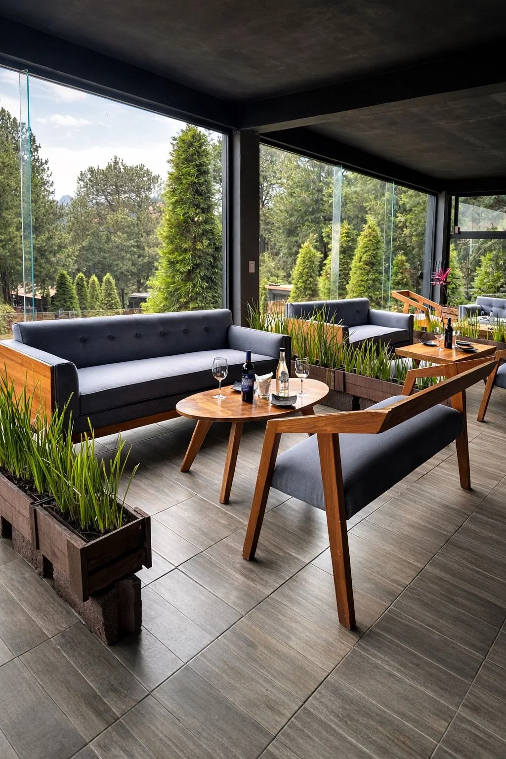 Interior de un salón con grandes ventanas de vidrio y decorado con lámparas colgantes. Hay personas, una de ellas sentada en un sillón y otra de pie, con árboles verdes visibles afuera.