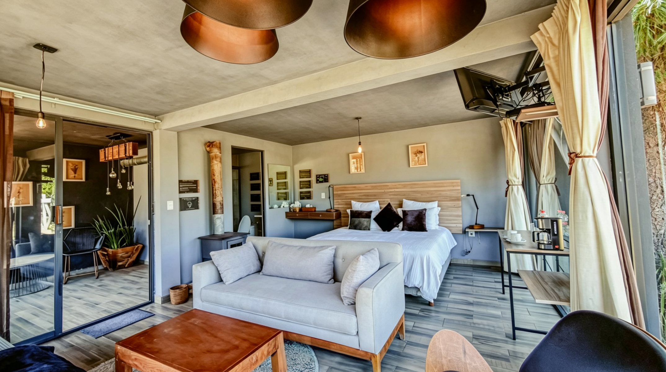 Habitación de hotel con cama grande, sofá gris, mesa de café de madera, cortinas claras y ventanales grandes con vista exterior, decorada con cuadros y plantas.