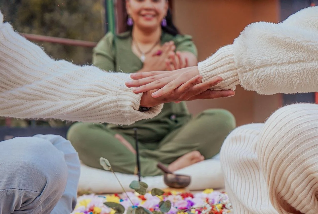 Dos personas se dan la mano en un ritual o ceremonia, con una mujer en el fondo que sonríe y observa, en un entorno al aire libre con flores y objetos ceremoniales en el suelo.